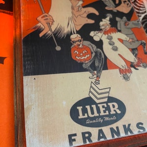 May include: A vintage Halloween party book cover with a witch, a clown, and a pumpkin. The book cover is framed in wood with a red border. The text on the cover reads "Party Book" and "WEENY-WITCH". The bottom of the cover has a black oval with the text "Luer Quality Meats" and "FRANKS".