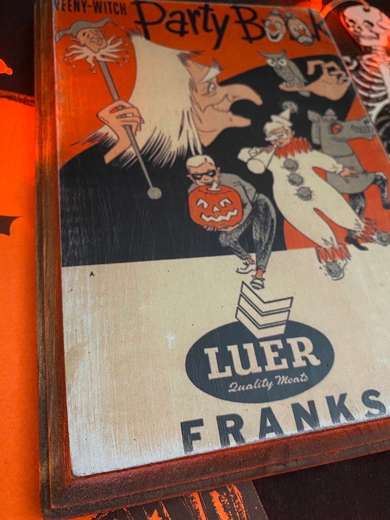 May include: A vintage Halloween party book cover with a witch, a clown, and a pumpkin. The book cover is framed in wood with a red border. The text on the cover reads "Party Book" and "WEENY-WITCH". The bottom of the cover has a black oval with the text "Luer Quality Meats" and "FRANKS".