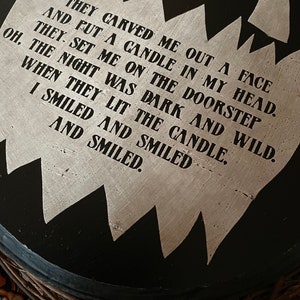Op de afbeelding: Een rond zwart houten bord met een gekarteld, lichtgekleurd midden met de tekst: THEY CARVED ME OUT A FACE AND PUT A CANDLE IN MY HEAD. THEY SET ME ON THE DOORSTEP OH, THE NIGHT WAS DARK AND WILD, WHEN THEY LIT THE CANDLE, I SMILED AND SMILED AND SMILED.