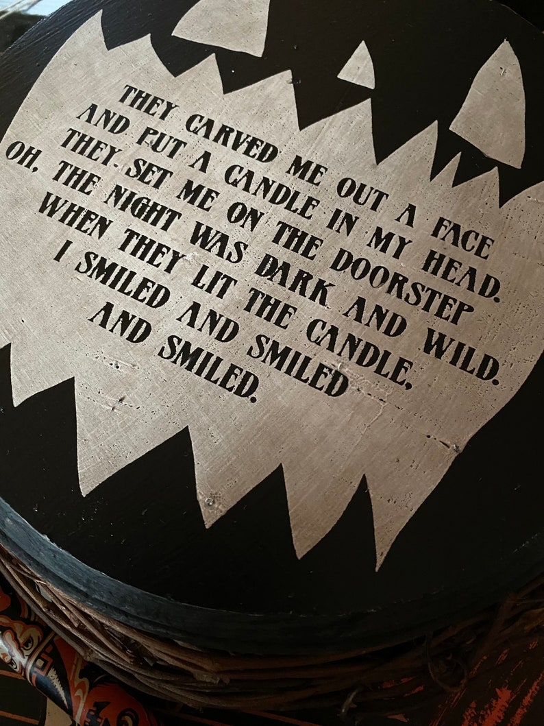 Op de afbeelding: Een rond zwart houten bord met een gekarteld, lichtgekleurd midden met de tekst: THEY CARVED ME OUT A FACE AND PUT A CANDLE IN MY HEAD. THEY SET ME ON THE DOORSTEP OH, THE NIGHT WAS DARK AND WILD, WHEN THEY LIT THE CANDLE, I SMILED AND SMILED AND SMILED.