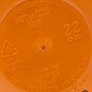 Peut inclure: Gros plan sur un mug isotherme orange. Le mug porte l'inscription "INSULATED HOT or COLD MUG" et "22 OZ.". Le bas du mug indique "ALPHA PRODUCTS ATLANTA, GA." et "MADE IN U.S.A."