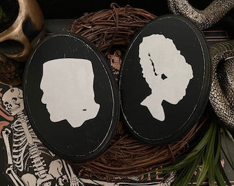 Puede incluir: Dos letreros circulares de madera negra con siluetas blancas de la cabeza y los hombros de una persona. El letrero de la izquierda presenta la silueta de una persona con una mandíbula cuadrada y cabello corto. El letrero de la derecha presenta la silueta de una persona con cabello largo y ondulado.