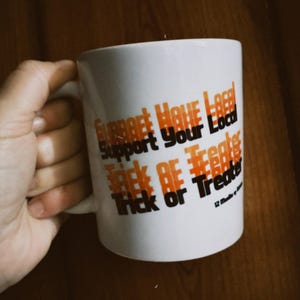 以下が含まれることがあります： オレンジと黒で「Support Your Local Trick or Treat」と書かれた白いセラミックマグ。マグには取っ手があり、手で持たれています。マグは標準サイズです。