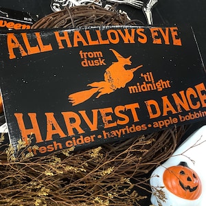Pode incluir: Uma placa de madeira preta com texto laranja e uma ilustração de bruxa. A placa diz "ALL HALLOWS EVE" e "HARVEST DANCE". Está colocada contra uma coroa de videira, com outras decorações de Halloween visíveis.