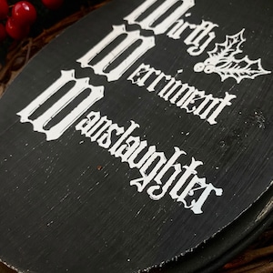 Puede incluir: Un letrero ovalado de madera negra con el texto blanco Mirth, Merriment, Manslaughter, acompa&ntilde;ado de una ilustraci&oacute;n de l&iacute;neas blancas de hojas y bayas de acebo. El letrero tiene un acabado pintado texturizado con pinceladas visibles.