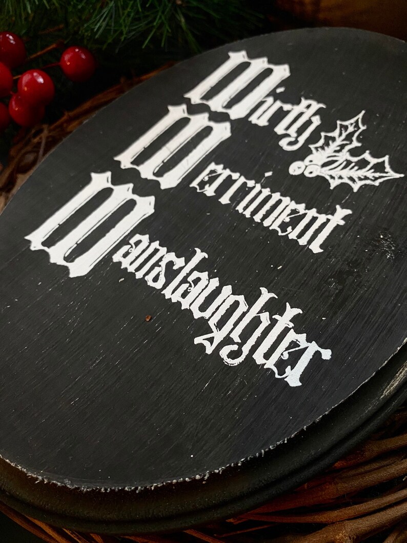 Puede incluir: Un letrero ovalado de madera negra con el texto blanco Mirth, Merriment, Manslaughter, acompa&ntilde;ado de una ilustraci&oacute;n de l&iacute;neas blancas de hojas y bayas de acebo. El letrero tiene un acabado pintado texturizado con pinceladas visibles.