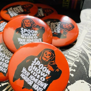 May include: Five orange round buttons with a black and white illustration of a hooded figure with a skull face. The text on the buttons reads "Spook Show Video Your Hometown Horror House".