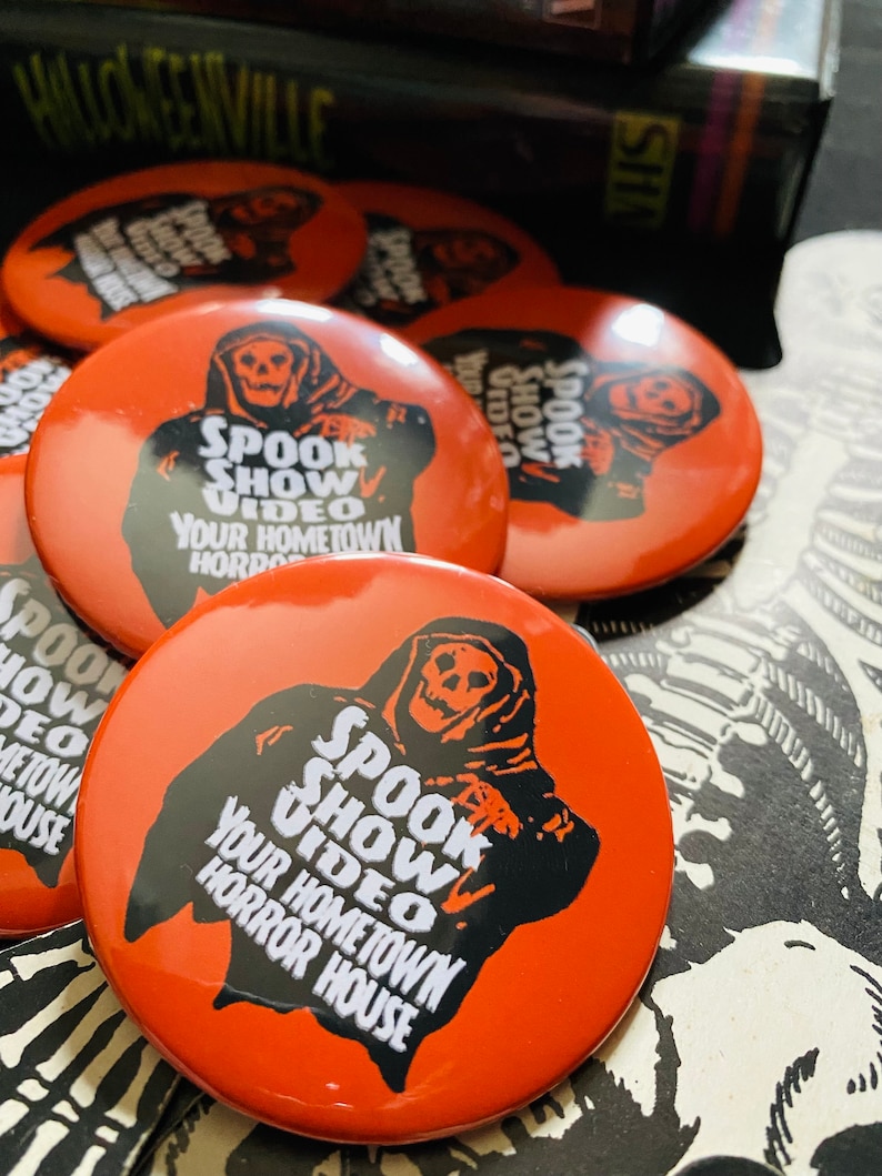 May include: Five orange round buttons with a black and white illustration of a hooded figure with a skull face. The text on the buttons reads "Spook Show Video Your Hometown Horror House".