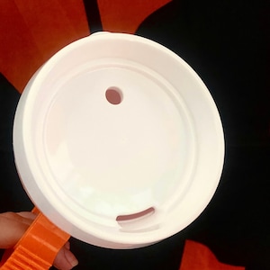 Peut inclure: Un couvercle de tasse en plastique blanc avec un petit trou et une ouverture incurv&eacute;e. Le couvercle est tenu par une poign&eacute;e orange. Le fond est noir et orange.