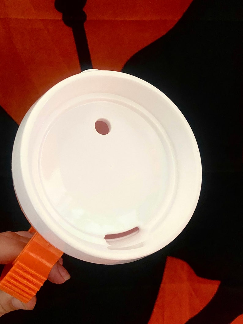 Peut inclure: Un couvercle de tasse en plastique blanc avec un petit trou et une ouverture incurv&eacute;e. Le couvercle est tenu par une poign&eacute;e orange. Le fond est noir et orange.