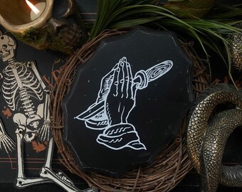 May include: Black and white illustration of a hand holding a knife, with the fingers in a praying position. The illustration is on a black circular wooden sign.