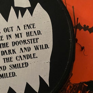 Op de afbeelding: Een rond zwart houten bord met een witte uitsparing in de vorm van een geest met zwarte tekst: CUT OUT A FACE, HOLE IN MY HEAD, ON THE DOORSTEP, DARK AND WILD, LIGHT THE CANDLE, AND SMILED, AND SMILED. Een oranje pompoenvormig bord is gedeeltelijk zichtbaar op de achtergrond.