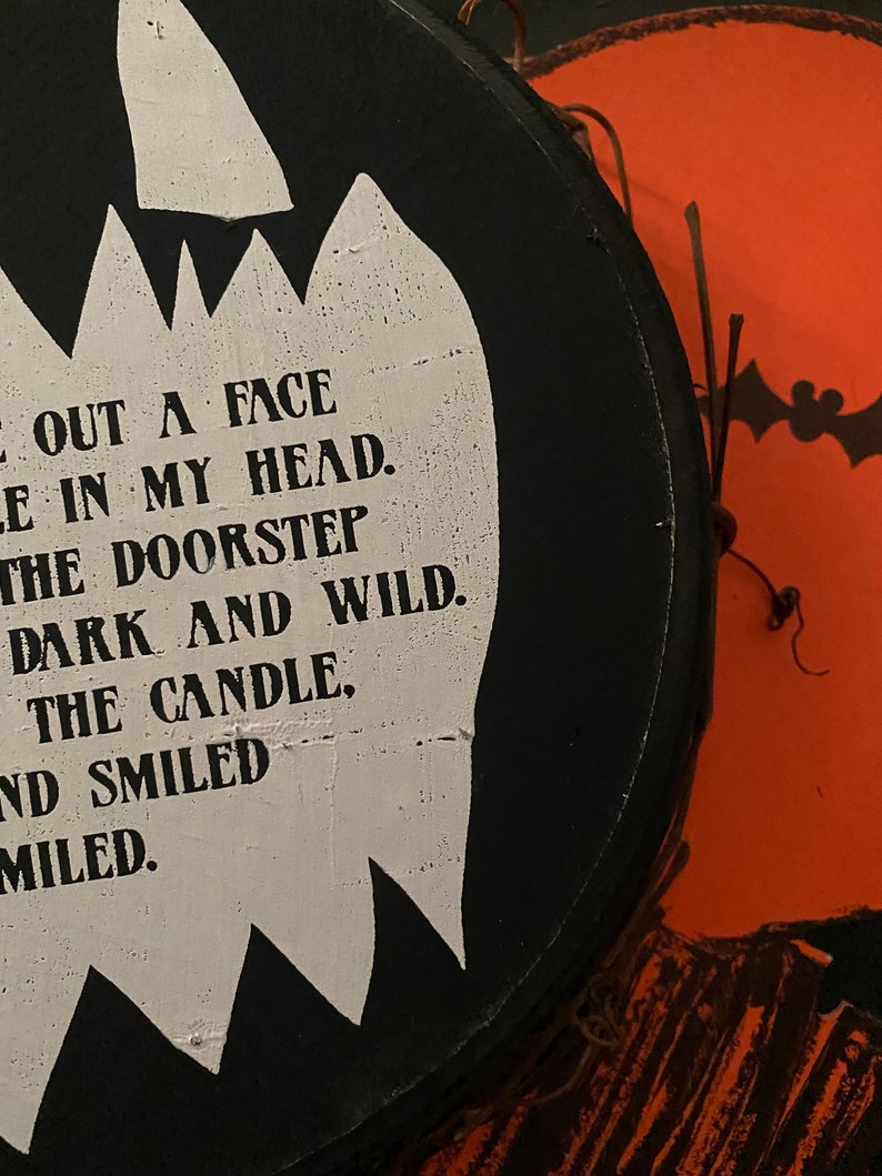 Op de afbeelding: Een rond zwart houten bord met een witte uitsparing in de vorm van een geest met zwarte tekst: CUT OUT A FACE, HOLE IN MY HEAD, ON THE DOORSTEP, DARK AND WILD, LIGHT THE CANDLE, AND SMILED, AND SMILED. Een oranje pompoenvormig bord is gedeeltelijk zichtbaar op de achtergrond.