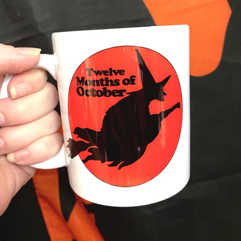 May include: White ceramic mug with a red circle and a black silhouette of a witch flying on a broomstick. The text "Twelve Months of October" is printed in black on the mug.