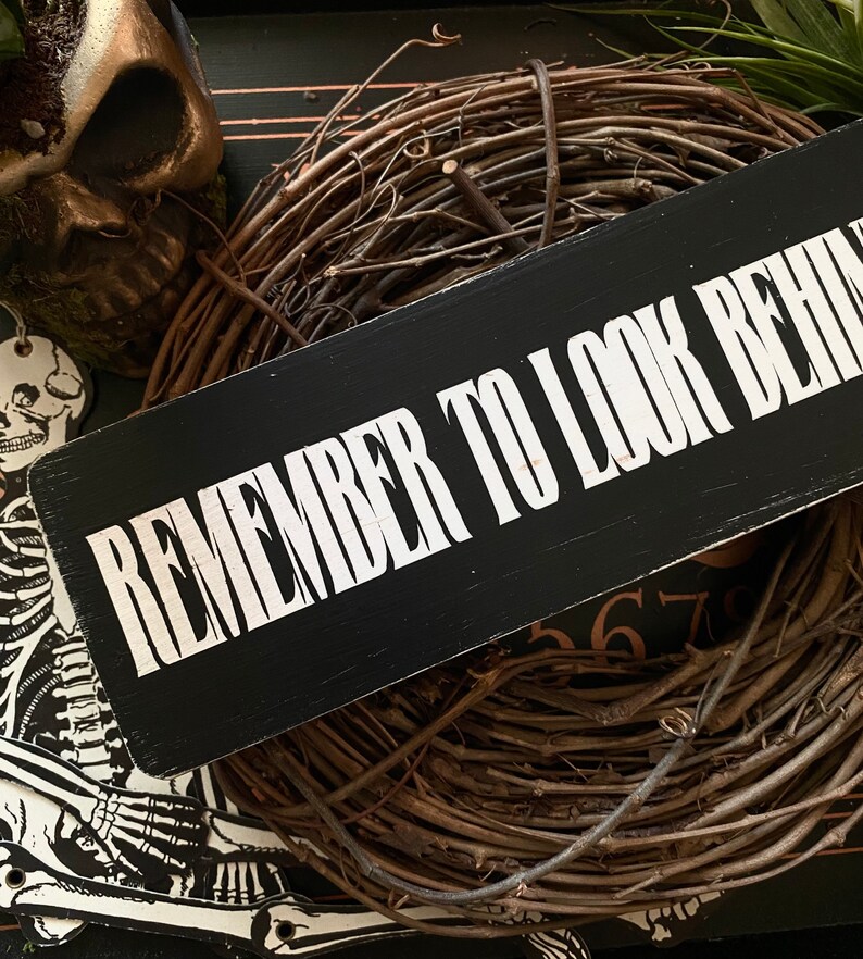 Może przedstawiać: Czarny drewniany znak z białym napisem "Remember to look behind you". Znak jest oparty o wieniec z brązowych gałązek.