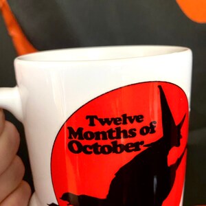 May include: White ceramic mug with a red circle and black silhouette of a witch flying. The circle has the text "Twelve Months of October" in black.