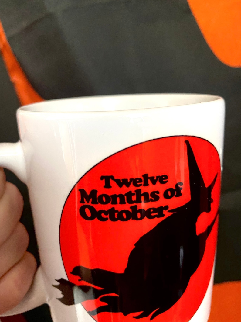 May include: White ceramic mug with a red circle and black silhouette of a witch flying. The circle has the text "Twelve Months of October" in black.