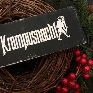 May include: Black wooden sign with white text that reads "Krampusnacht" and a white silhouette of a Krampus figure. The sign is leaning against a brown grapevine wreath with red berries.