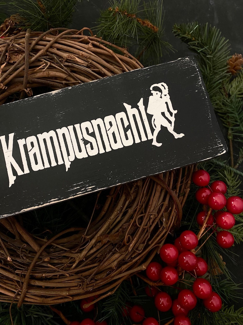 May include: Black wooden sign with white text that reads "Krampusnacht" and a white silhouette of a Krampus figure. The sign is leaning against a brown grapevine wreath with red berries.