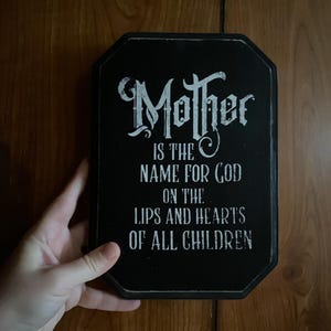 Może przedstawiać: Czarny drewniany znak z cytatem "Mother is the name for God on the lips and hearts of all children" w białych literach. Znak ma ośmiokątny kształt i jest trzymany w dłoni.