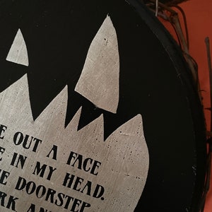 Op de afbeelding: Een zwarte, ronde houten plank met zilveren tekst en een gekarteld ontwerp. De tekst luidt "E OUT A FACE IN MY HEAD. E DOORSTEP DARK AND WILD. CANDLE". Een decoratieve takkenrand is zichtbaar.