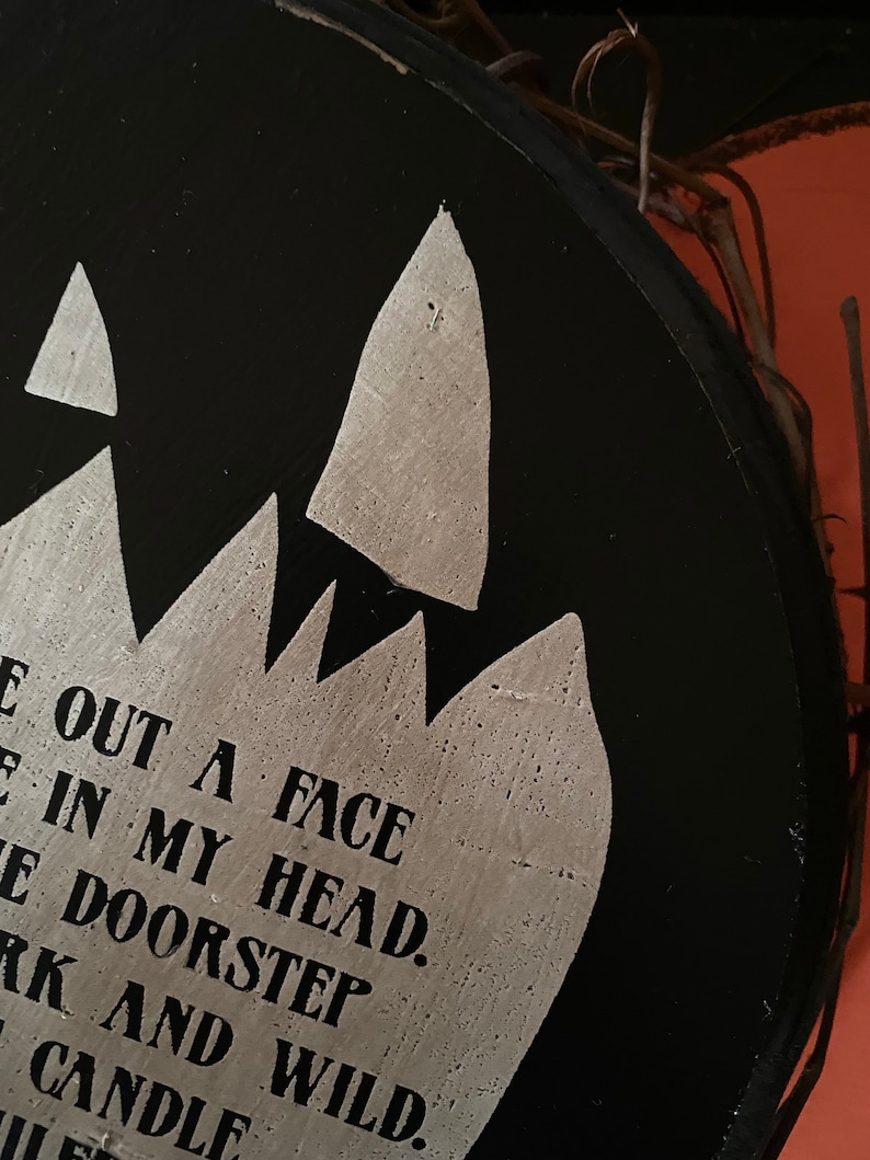 Op de afbeelding: Een zwarte, ronde houten plank met zilveren tekst en een gekarteld ontwerp. De tekst luidt "E OUT A FACE IN MY HEAD. E DOORSTEP DARK AND WILD. CANDLE". Een decoratieve takkenrand is zichtbaar.