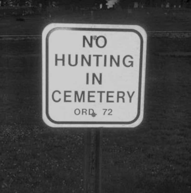 Op de afbeelding: Een zwart-wit bord met de tekst "NO HUNTING IN CEMETERY ORD. 72".