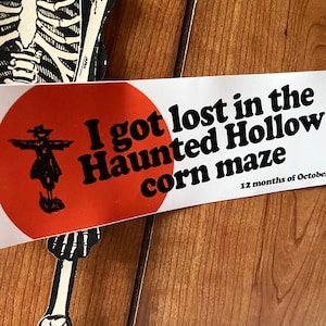 May include: A Halloween-themed sign with the text "I got lost in the Haunted Hollow corn maze" on a white background with an orange circle and a scarecrow graphic. A black and white skeleton decoration is in the image.