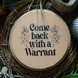 Może przedstawiać: Okrągły drewniany znak z beżowym tłem i czarnym tekstem, który brzmi "Come back with a warrant". Znak jest otoczony wieńcem z brązowych gałązek i zieloną rośliną.