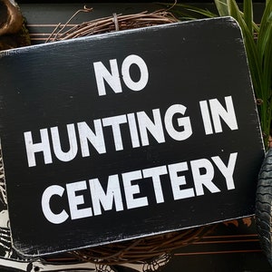 Op de afbeelding: Zwart houten bord met witte tekst die "NO HUNTING IN CEMETERY" luidt.