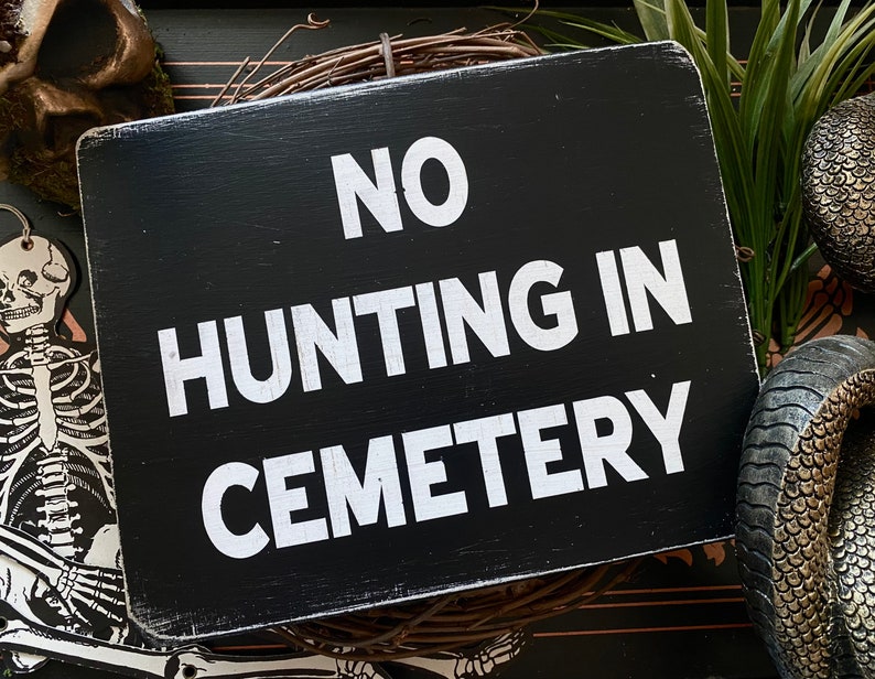 Op de afbeelding: Zwart houten bord met witte tekst die "NO HUNTING IN CEMETERY" luidt.