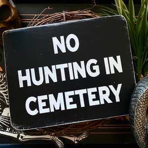 Op de afbeelding: Een zwart houten bord met witte tekst die "NO HUNTING IN CEMETERY" luidt. Het bord is omringd door een skelet, een schedel en een slang.