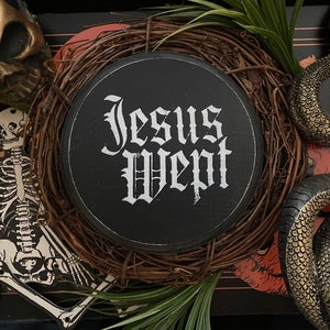 May include: A black circular sign with the words "Jesus Wept" written in white gothic font. The sign is surrounded by a wreath of brown twigs and vines. There is a gold skeleton and a gold snake in the background.