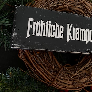 May include: A black wooden sign with white lettering that reads "Fr&ouml;hliche Krampus". The sign is hanging on a brown grapevine wreath decorated with red berries.