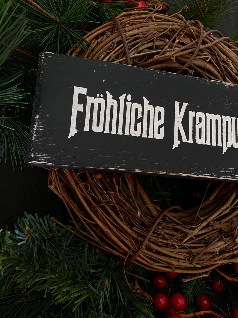 May include: A black wooden sign with white lettering that reads "Fr&ouml;hliche Krampus". The sign is hanging on a brown grapevine wreath decorated with red berries.