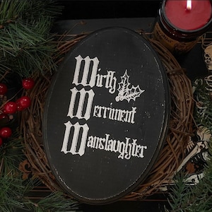 Puede incluir: Una placa ovalada de madera negra con letras g&oacute;ticas blancas que dicen Mirth, Merriment, Manslaughter, acompa&ntilde;ada de una peque&ntilde;a ilustraci&oacute;n blanca de hojas y bayas de acebo centrada en la superficie.