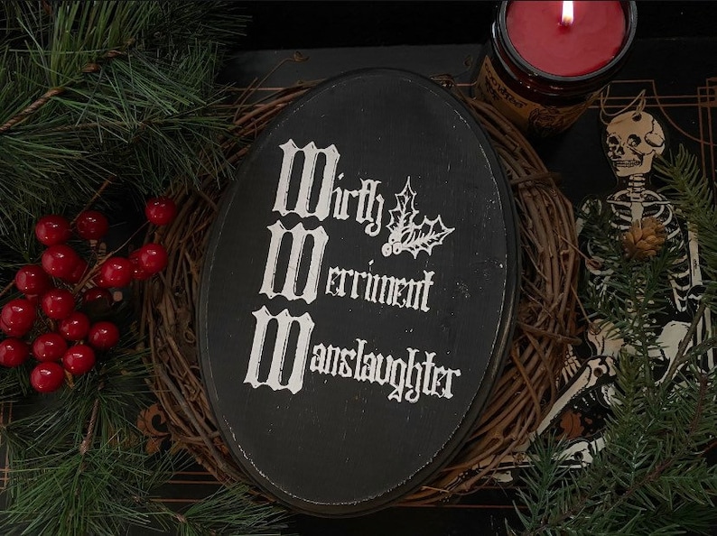 Puede incluir: Una placa ovalada de madera negra con letras g&oacute;ticas blancas que dicen Mirth, Merriment, Manslaughter, acompa&ntilde;ada de una peque&ntilde;a ilustraci&oacute;n blanca de hojas y bayas de acebo centrada en la superficie.