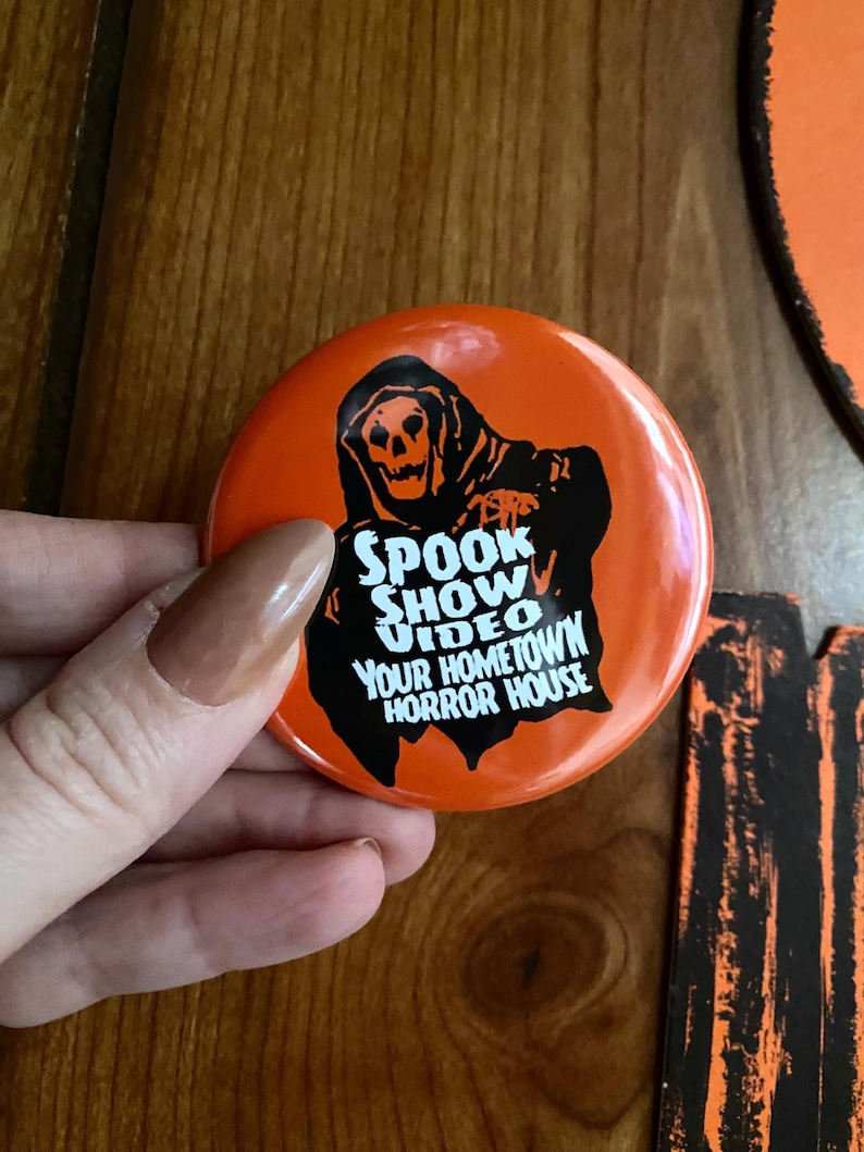 May include: Orange round button featuring a black and white image of a skeleton in a hooded robe. The text on the button reads "Spook Show Video Your Hometown Horror House".