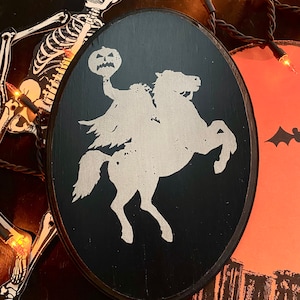 Op de afbeelding: Een zwart ovaal houten bord met een wit silhouet van een koploze ruiter die een paard berijdt en een jack-o-lantern vasthoudt.