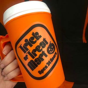Peut inclure: Mug d'Halloween en plastique orange avec un couvercle et une poign&eacute;e blancs. Le mug pr&eacute;sente une &eacute;tiquette noire et orange avec le texte "Trick or Treat Mart" et un dessin de citrouille. Le texte "Open 24 Hours" est &eacute;galement sur l'&eacute;tiquette.