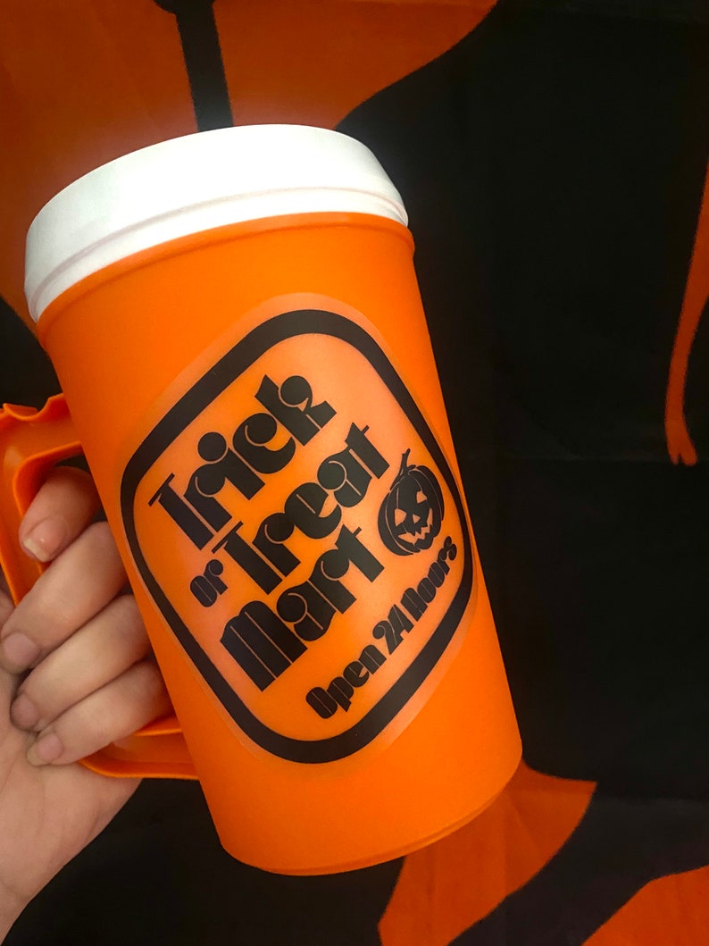 Peut inclure: Mug d'Halloween en plastique orange avec un couvercle et une poign&eacute;e blancs. Le mug pr&eacute;sente une &eacute;tiquette noire et orange avec le texte "Trick or Treat Mart" et un dessin de citrouille. Le texte "Open 24 Hours" est &eacute;galement sur l'&eacute;tiquette.