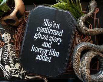 Puede incluir: Un letrero de madera negro en forma de ataúd con texto blanco: "She's a confirmed ghost story and horror film addict". El letrero está rodeado por una corona decorativa y decoraciones temáticas de Halloween.