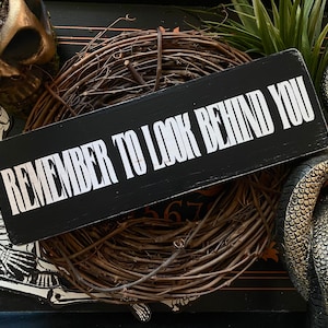 Może przedstawiać: Czarny drewniany znak z białym tekstem, kt&oacute;ry brzmi "Remember to look behind you". Znak jest otoczony wieńcem z gałązek i innymi dekoracjami halloweenowymi.
