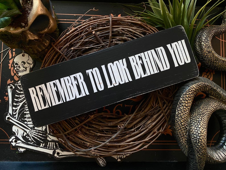 Może przedstawiać: Czarny drewniany znak z białym tekstem, kt&oacute;ry brzmi "Remember to look behind you". Znak jest otoczony wieńcem z gałązek i innymi dekoracjami halloweenowymi.
