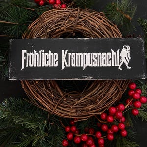May include: A black wooden sign with white text that reads "Fr&ouml;hliche Krampusnacht!" with a white silhouette of a Krampus figure on the right side. The sign is hanging on a brown grapevine wreath decorated with red berries and evergreen branches.
