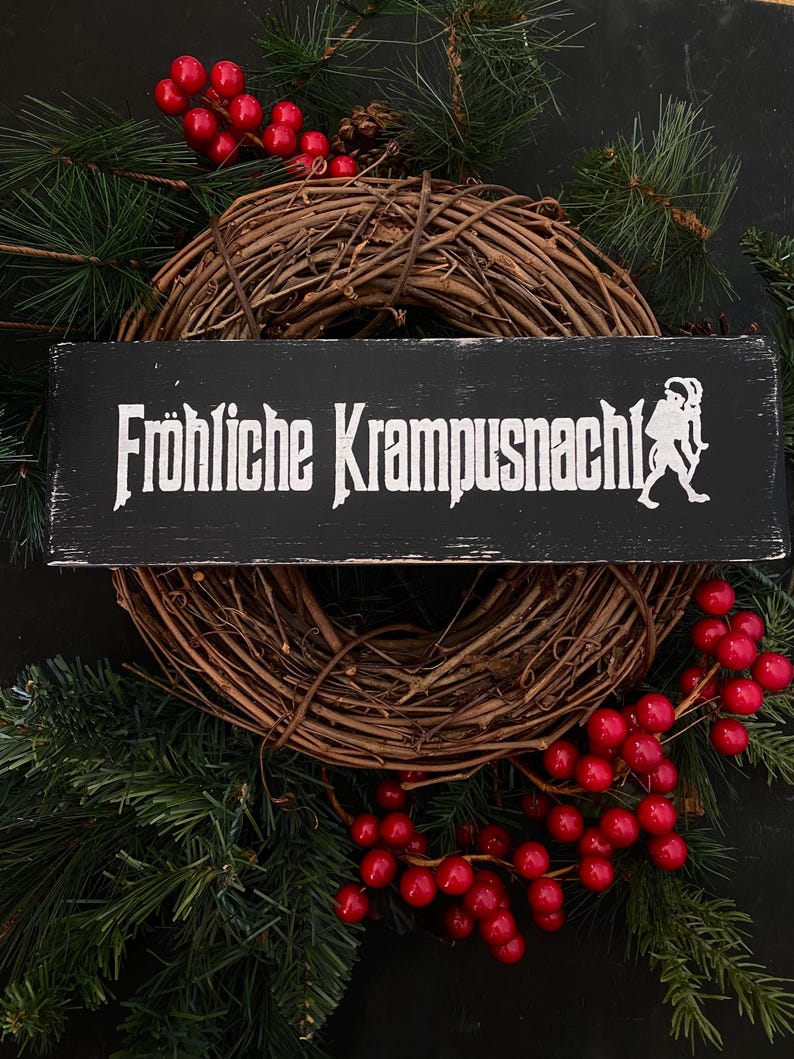 May include: A black wooden sign with white text that reads "Fr&ouml;hliche Krampusnacht!" with a white silhouette of a Krampus figure on the right side. The sign is hanging on a brown grapevine wreath decorated with red berries and evergreen branches.