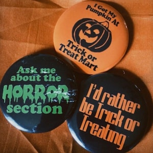 Puede incluir: Tres botones con temática de Halloween. Un botón naranja con una calabaza y el texto "I Got My Pumpkin At Trick or Treat Mart". Los otros dos son negros, con las frases "Ask me about the HORROR section" y "I'd rather be Trick or Treating."