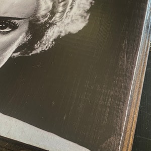 Puede incluir: Primer plano en blanco y negro de la cara de una mujer con un peinado que recuerda al monstruo de Frankenstein. La imagen est&aacute; impresa en una placa de madera.