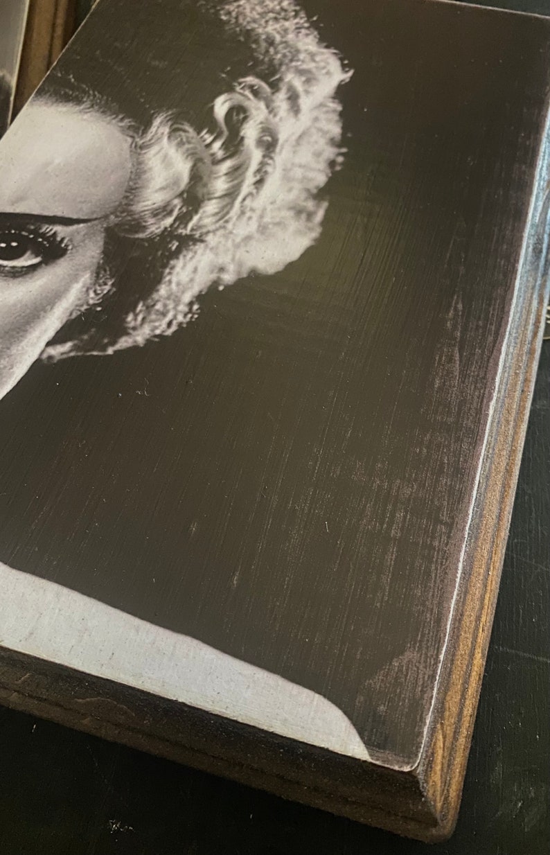 Puede incluir: Primer plano en blanco y negro de la cara de una mujer con un peinado que recuerda al monstruo de Frankenstein. La imagen est&aacute; impresa en una placa de madera.