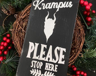 Può includere: Un cartello in legno nero con testo bianco che recita "Krampus Please Stop Here". Il cartello presenta una silhouette bianca di Krampus con corna e barba.